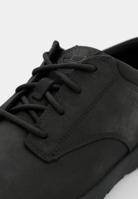 Zapatilla de cuero negro con acabado mate texturizado, que presenta cordones negros planos, suela de goma y un logotipo elevado en la lengüeta.