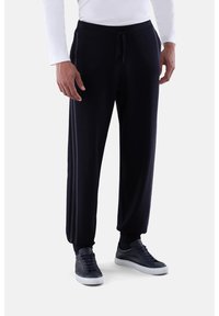 Mann in schwarzen Jogginghosen mit seitlichen Streifen, schwarzen Sneakers und einem weißen Langarmshirt, steht vor einem schlichten hellen Hintergrund.