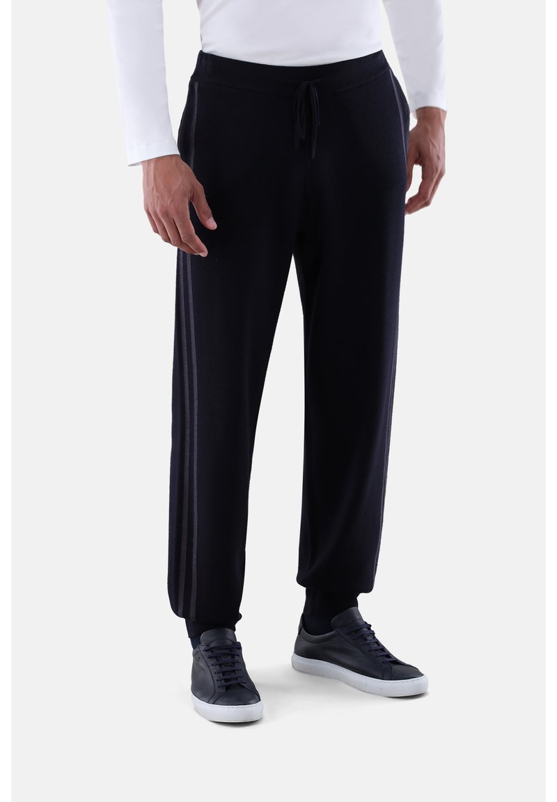 Mann in schwarzen Jogginghosen mit seitlichen Streifen, schwarzen Sneakers und einem weißen Langarmshirt, steht vor einem schlichten hellen Hintergrund.