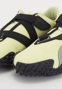 Chaussures de sport jaune clair avec des sangles noires croisées, semelles noires texturées avec des picots en caoutchouc, et logo Puma sur la sangle.