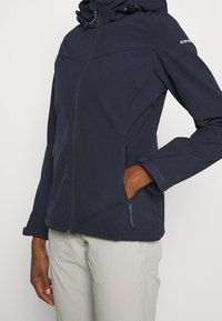 Granatowa kurtka softshell z zamkiem błyskawicznym, regulowanym kapturem i bocznymi kieszeniami. Materiał jest gładki o matowym wykończeniu i minimalnych szwach.