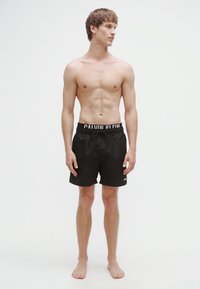 Μαύρα μαγιό σορτς με κορδόνι στη μέση και ζώνη με λογότυπο. Το μοντέλο στέκεται ξυπόλυτο, παρουσιάζοντας έναν απλό αθλητικό σχεδιασμό χωρίς σχέδια.