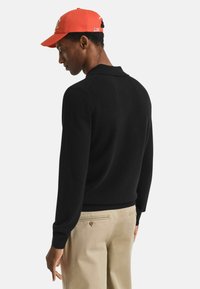 Zwarte gebreide trui met lange mouwen, kraag, gecombineerd met lichtbeige chinos en een oranje pet met een logo. Achteraanzicht.