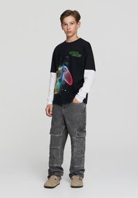 Czarny t-shirt z grafiką w multikolorowe kontrolery do gier i zielonym napisem, nałożony na białe długie rękawy. Szare spodnie cargo i beżowe buty.