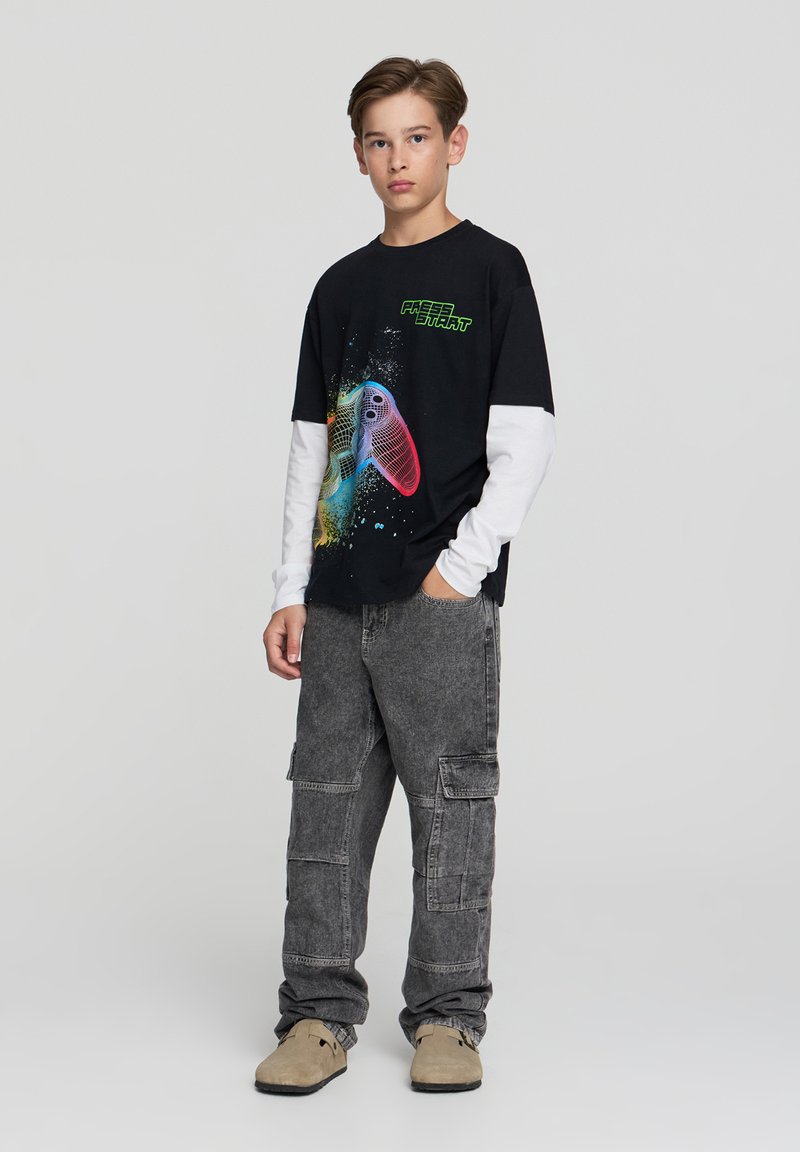 Czarny t-shirt z grafiką w multikolorowe kontrolery do gier i zielonym napisem, nałożony na białe długie rękawy. Szare spodnie cargo i beżowe buty.