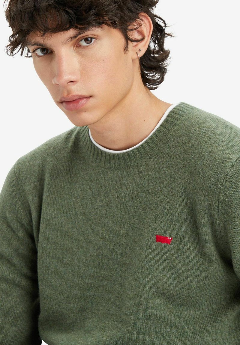 Maglione verde in lana con scollo rotondo, polsini a coste e una piccola toppa rossa sul lato sinistro del petto. Texture liscia e design aderente.
