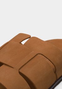 Εγγύς εικόνα καφέ σουέντ σανδάλι με φαρδιές, διασταυρούμενες λουριά και ανοιχτό σχέδιο μύτης πάνω σε ανοιχτό φωτεινό φόντο.