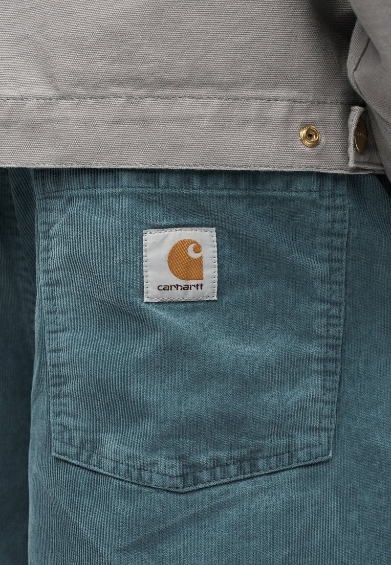 Gros plan sur la poche d'un pantalon en velours côtelé bleu sarcelle avec une étiquette Carhartt blanche et orange, associé à une veste gris clair avec des boutons-pression métalliques.