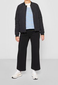 Svart vadderad bomberjacka över en vit och blå randig topp, matchad med svarta vida byxor och ljusa sneakers med ett vågmönster.