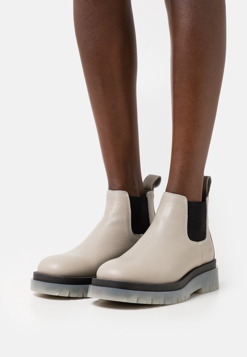 Bottines beige clair avec des panneaux latéraux élastiques noirs et des semelles crantées translucides portées sur des jambes à la peau foncée sur fond blanc.