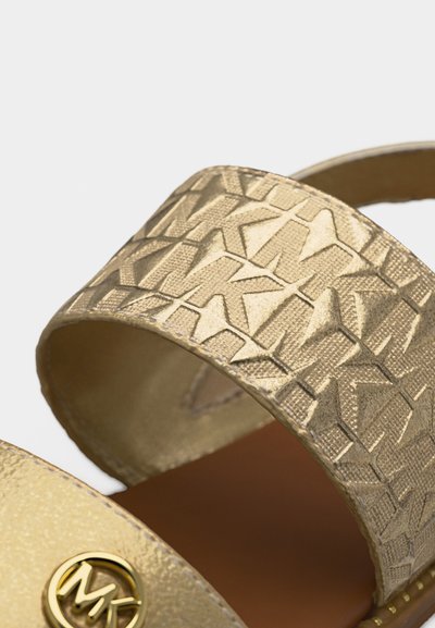 Λουράκι χρυσού σανδαλιού με ανάγλυφο επαναλαμβανόμενο μοτίβο λογότυπου "MK" και στρογγυλή μεταλλική εμβληματική λεπτομέρεια "MK" κοντά στην περιοχή των δακτύλων σε καφέ πάτο.
