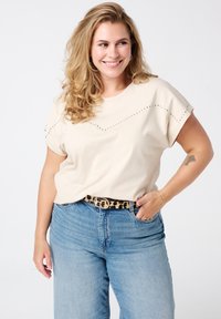 T-shirt beige à manches courtes avec un design clouté sur la poitrine, associé à un jean taille haute bleu clair et une ceinture à imprimé léopard.