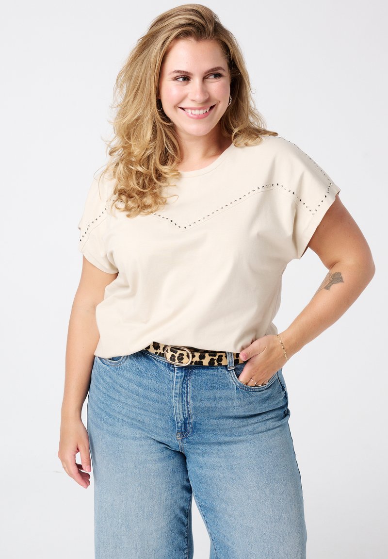 T-shirt beige à manches courtes avec un design clouté sur la poitrine, associé à un jean taille haute bleu clair et une ceinture à imprimé léopard.