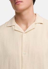 Camisa de manga corta beige con un tejido texturizado, abotonada en el cuello, que presenta un sutil logo bordado en el lado izquierdo.