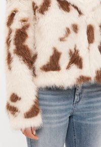 Casaco de pelúcia com uma base creme e padrão de vaca castanho, com uma textura suave e fofinha e mangas largas, combinado com jeans azuis.