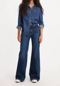 Femme portant une chemise en denim bleu foncé boutonnée aux manches retroussées et un jean flare taille haute associé à des baskets blanches.