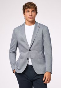 Blazer grigio chiaro con rever a notches, due tasche frontali e due bottoni neri. Indossato sopra una maglietta bianca, in tessuto strutturato con un motivo sottile.