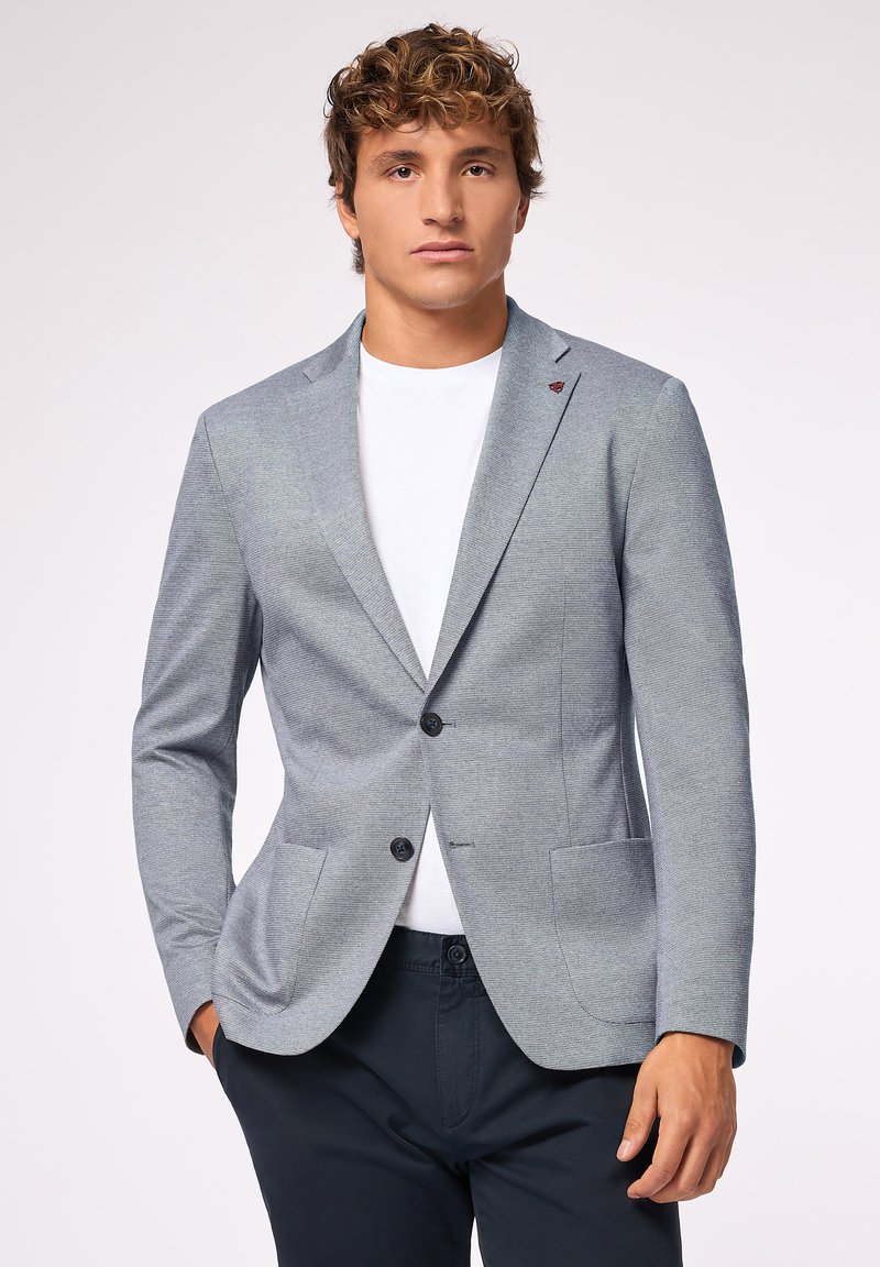 Blazer grigio chiaro con rever a notches, due tasche frontali e due bottoni neri. Indossato sopra una maglietta bianca, in tessuto strutturato con un motivo sottile.