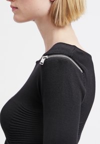 Femme vue de profil portant un pull en maille noire avec un détail de fermeture éclair argentée le long de la couture de l'épaule, cheveux blonds courts.