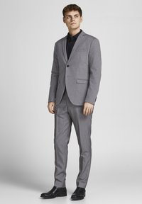 Costume gris sur mesure avec un blazer structuré et un pantalon droit, associé à une chemise noire et des bottes noires brillantes.