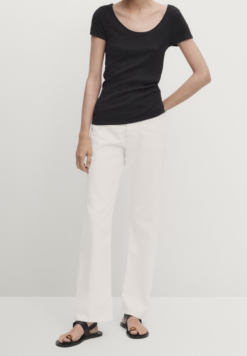 Femme portant un haut noir ajusté à manches courtes, un pantalon blanc droit et des sandales plates noires avec un détail anneau doré, debout avec une main sur la hanche.