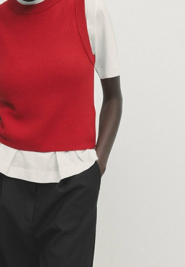 Red ribbed knit sleeveless top layered over a white short-sleeve shirt, paired with black trousers, showcasing a relaxed fit.