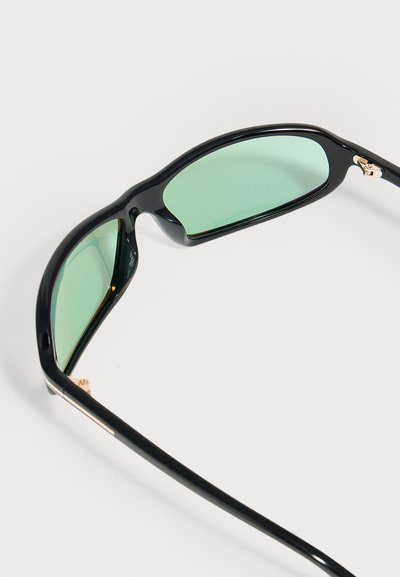 Lunettes de soleil à monture noire avec des verres teintés verts reposant sur une surface blanche, montrées sous un angle mettant en valeur la monture et les branches.