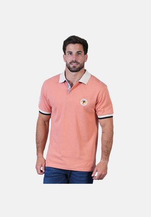 Homme portant un polo rose saumon avec col blanc et poignets de manches à rayures noires, associé à un jean bleu foncé, debout devant un fond blanc.
