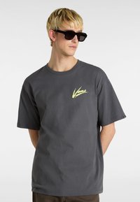 Camiseta de manga corta gris con un logo amarillo de Vans en el lado izquierdo del pecho y un corte relajado. El modelo lleva gafas de sol negras y las manos en los bolsillos.