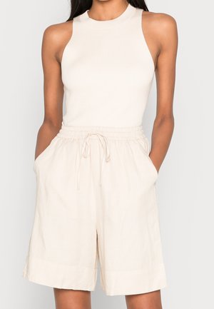 Woman wearing sleeveless ribbed cream top and high-waisted cream drawstring shorts with hands in pockets.