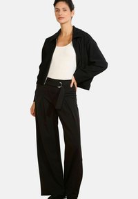 Čierna skrátená bunda s golierom a predným zipsom, doplnená o čierne široké nohavice s vysokým pásom, opaskom a skladanými detailmi.
