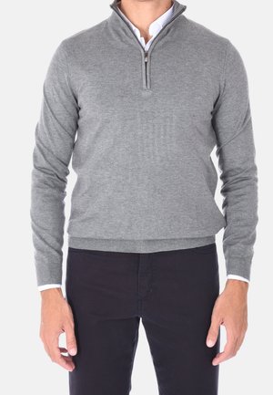 Hombre con un suéter de cremallera corta gris claro sobre una camisa blanca con cuello, combinado con pantalones negros, frente a un fondo liso.