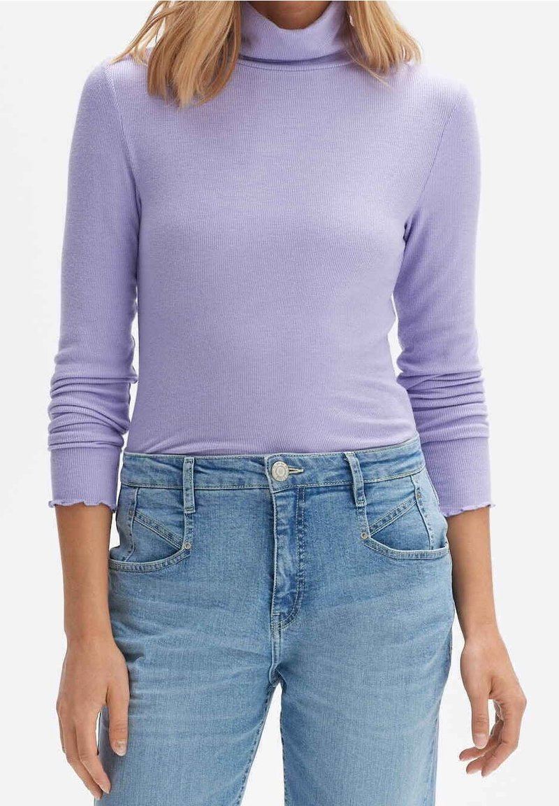 Frau in einem figurbetonten lila Ripp-Turtleneck mit 3/4-Ärmeln und hellblauen, hochtaillierten Jeans mit vorderen Taschen.