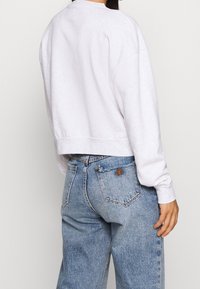 Valkoinen lyhyt kiristysvetoketjuhuppari, jossa on pyöreä kaula-aukko ja roikkuvat olkapäät, yhdistettynä vaaleansinisisiin korkeavyötäröisiin farkkuihin, joissa on kolmion muotoinen logo-ompelu.