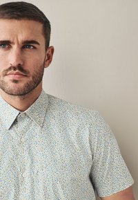 Camicia a maniche corte azzurro chiaro con motivo floreale giallo e blu scuro. Presenta un colletto classico e chiusura con bottoni.