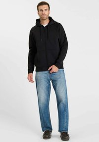 Zwarte hoodie met ritssluiting en een buidelzak, gecombineerd met lichtblauwe jeans en donkere schoenen. Heeft een gevoerde capuchon en geribbelde manchetten.