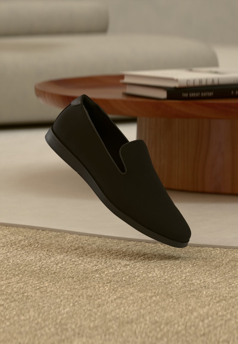 Chaussure noire sans lacets flottant au-dessus d'un tapis beige tissé, avec une table basse ronde en bois et des livres empilés en arrière-plan flou.