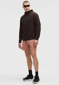 Barna vízálló dzseki kapucnival, világos rózsaszín rövidnadrág felett viselve, fekete sportcipőkkel és napszemüveggel párosítva. Sima textúra, minimalista design.