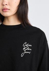 Crna pamučna majica s bijelim ručno napisanom logotipom "Calvin Klein Jeans" na lijevoj strani grudnog područja. Jednostavna rundi vrat.
