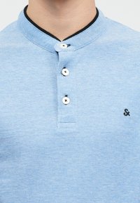 Vaaleansininen poolopaita, jossa on teksturoidut kankaat, musta kauluskoristelu, kolme nappia ja pieni musta logo vasemmassa rinnassa.