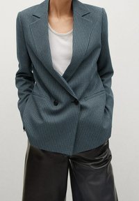 Veste croisée à carreaux bleu sarcelle avec revers crantés et deux poches avant, associée à un t-shirt blanc à col rond et un pantalon en cuir noir.