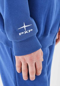 Blauwe sweatshirt met ribbels aan de manchetten, met een opvallend wit "PXP" logo en steraccent op de linker mouw. Soepele textuur, casual ontwerp.