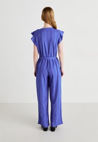 Paarse jumpsuit met zigzagpatroon, korte gerimpelde mouwen, een ceintuur in de taille en een wijd uitlopende broekspijp, van achteraf gezien tegen een neutrale achtergrond.