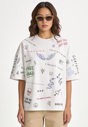 Frau trägt beigefarbene Sonnenbrille und ein weißes, übergroßes T-Shirt, bedeckt mit bunten handgeschriebenen Phrasen und kleinen Skizzen.