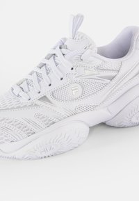 Zapatilla deportiva blanca con una parte superior de malla y superposiciones texturizadas, cordones planos y suela de goma con un dibujo en relieve. Logotipo de Fila en el lateral.