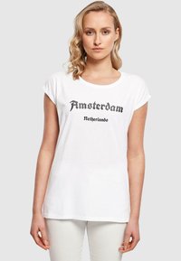 Valkoinen lyhythihainen t-paita, jossa on mustalla painettuna teksti "Amsterdam Alankomaat". T-paidassa on rento malli ja pyöreä pääntie.