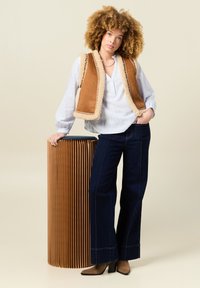 Gilet marrone con bordo in pelliccia, camicia bianca, jeans blu a gamba larga e stivaletti con tacco color beige. In piedi accanto a un oggetto cilindrico con righe.