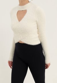 Maglione bianco morbido con collo alto e apertura a chiave, caratterizzato da un design avvolgente e maniche lunghe, abbinato a leggings neri a costine.