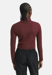 Person mit geflochtenem Haar, die ein langärmliges bordeauxfarbenes Shirt und schwarze Hosen trägt, steht mit dem Rücken zu einem schlichten Hintergrund.
