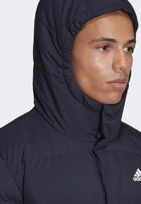 Marinblå puffjacka med ett kviltat mönster och en justerbar huva. Har en framzipper och en framträdande vit Adidas-logga.